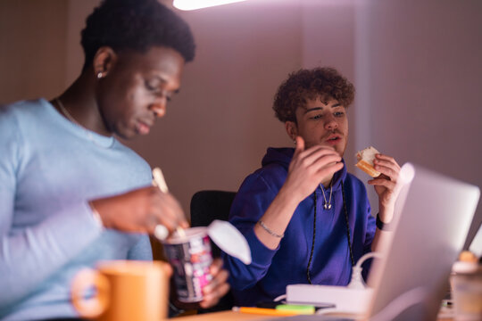 Two young men sit at a desk in a dimly lit workspace, one eating noodles with chopsticks and the other holding a sandwich while engaged in conversation.