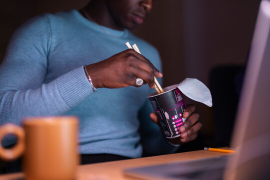 A man in a blue sweater is using chopsticks to eat instant noodles from a cup while working on his laptop at a dimly lit desk with a warm, ambient light.