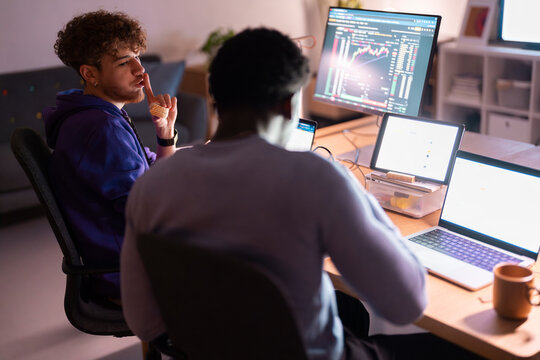 A young man in a blue hoodie eats a sandwich while another man works at a desk with multiple screens displaying stock market data and digital charts.