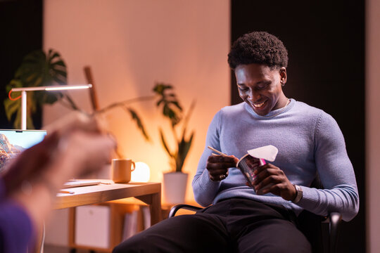 A young man in a blue sweater is laughing while eating instant noodles with chopsticks in a warmly lit workspace with a laptop and coffee mug nearby.
