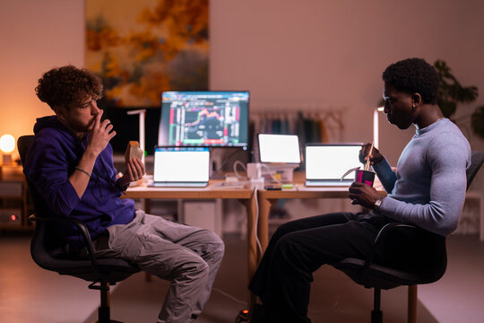 Two young men sit in a modern workspace with multiple screens displaying stock market data, one eating a sandwich and the other using chopsticks to eat instant noodles.