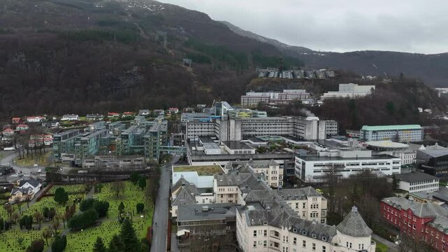 Haukeland Hospital and Helse Bergen in Norway with a graveyard on the side and mountain backdrop. Slow aerial rise on an overcast day