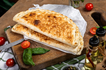 A golden-brown calzone rests on a wooden cutting board, surrounded by fresh basil leaves, cherry tomatoes, and a glass jar of mixed spices. A bottle of olive oil and a small knife are nearby	