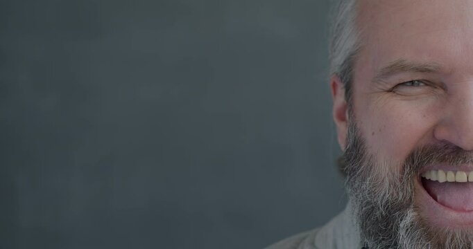 Close-up slow motion portrait of mature man half face laughing looking at camera on gray color background. People and good mood concept.