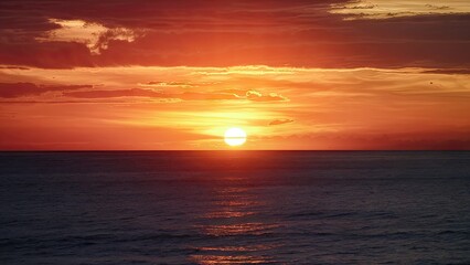 Naklejka premium Vibrant sunset casting warm orange and red hues over calm ocean waves reflecting the colorful sky at horizon line during serene evening.