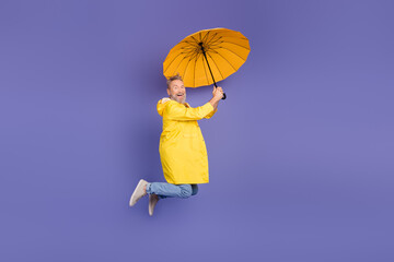 Joyful mature man in vibrant yellow raincoat jumping against a purple background while holding a...
