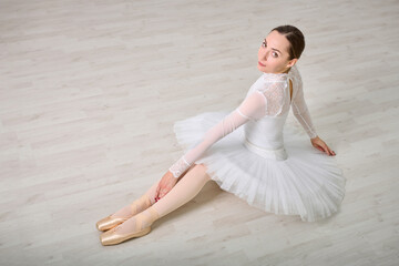 Young female ballerina with brown hair in white tutu, seated elegantly on wooden floor. Soft lighting enhances her poised expression and delicate pose, casting gentle shadows © TRAVELARIUM