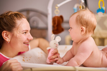 Smiling adult mother with brown hair plays with joyful baby girl on changing table. Soft lighting highlights calm, warm nursery ambiance, and colorful toy