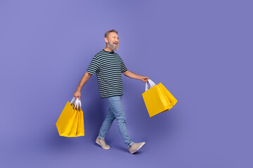 Smiling mature man with yellow shopping bags walking against purple backdrop, expressing happiness,...