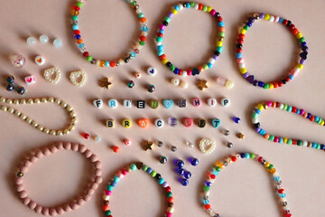 Friendship bracelet spelled with letter beads. Colorful handmade bracelets and various beads. Flat lay, pink background.