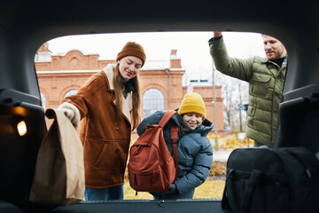 Family members loading luggage into car trunk during winter, standing near brick building with...