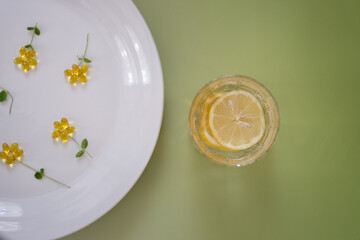 A top-down view of a still life composition with vitamin D capsules arranged as flowers on a white plate, accompanied by a glass of lemon water. 