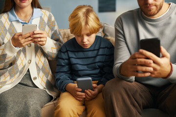 Family gathered on couch, engrossed in using smartphones and spending time together, child sitting...