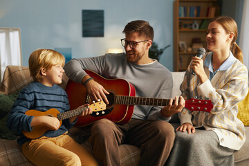 Family enjoying music session with father playing guitar, son playing ukulele, and daughter singing, all sitting on comfortable sofa, creating joyful atmosphere