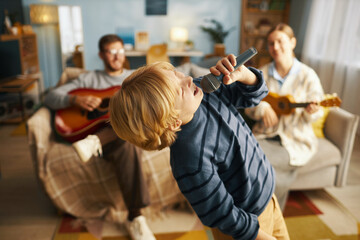 Blond child passionately singing with microphone while family guitarist and ukulele player strumming in cozy living room setting