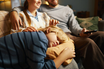 Family sitting on couch enjoying movie night together while young child naps on mothers lap parents...