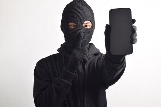 Mysterious man wearing black hoodie and mask doing hacking activity on smartphone, putting finger to lips, making silent sign, isolated on white background.