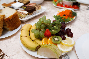 In the foreground is a plate of sliced ​​fruit: bananas, green grapes, strawberries, kiwis, oranges, lemons and pears.