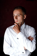 a man in a white shirt and dark trousers. He stands against a dark wall and looks to the side. He has short hair and a serious expression on his face.