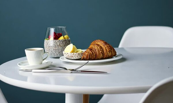 A beautifully arranged breakfast setting on a white table, featuring a golden-brown croissant on a white plate, accompanied by a small bowl of butter. In the background, a speckled glass container hol