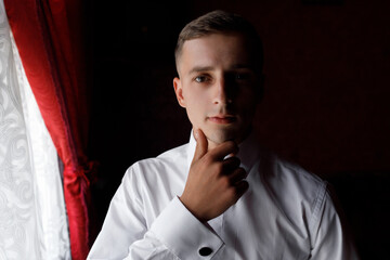 a man with short dark hair and fair skin. He has brown eyes and neat facial features. He is wearing a white shirt and looking straight into the camera. 
