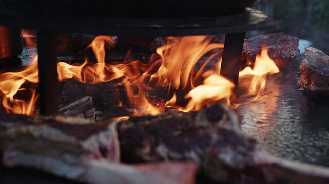 Large cuts of meat grill above roaring flames in a rustic outdoor kitchen. Smoke billows as the meat sizzles, creating an inviting atmosphere of cooking and camaraderie.
