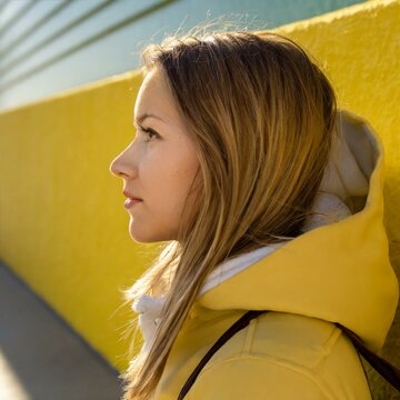 Eine blonde junge Frau in gelber Jacke vor einer gelben Wand schaut nachdenklich in die Ferne.
