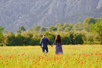A man and a woman walk hand in hand through a field with red poppies against the backdrop of beautiful mountains on a sunny day