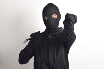Thief wearing black jacket and mask holding crowbar, posing pointing at camera, isolated on white...