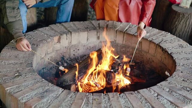 A mans hand holds a wooden skewer with a marshmallow over an open fire in a rustic fire pit. The warm flames and crackling wood create a peaceful and cozy outdoor moment. Perfect for themes of camping