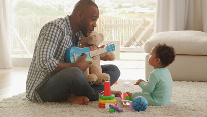 Father And Young Son Playing Music On Toy Guitar Or Ukulele At Home Together