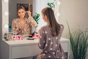 Young woman in polka dot pajama applying makeup at home in morning light, sitting at vanity in cozy...