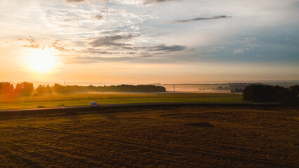 Fototapeta premium dawn in a wheat field, a car is driving along the highway, fog is creeping on the horizon