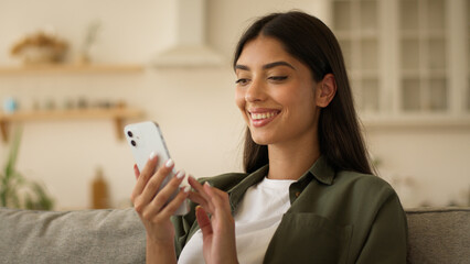 Happy smiling Indian woman Arabian girl female Muslim lady at home couch sofa kitchen shopping online mobile phone order food delivery scrolling smartphone social media app smile internet using net