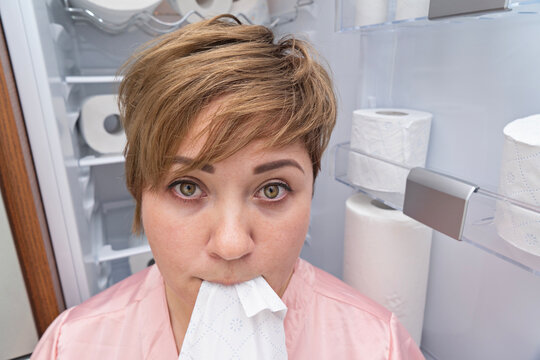 Pregnant woman with short hair and confused expression holds toilet paper in her mouth, standing in front of fully stocked toilet paper shelf. Non-food items eating concept