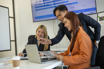 Three people are sitting at a table with a laptop open. One of them is laughing while the other two look on. The man in the middle is giving a presentation to the other two
