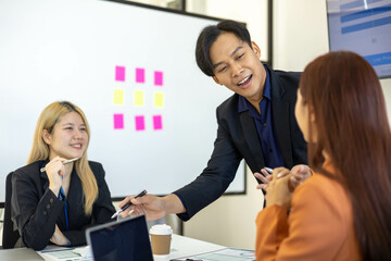 Three people are sitting at a table with a laptop open. One of them is laughing while the other two look on. The man in the middle is giving a presentation to the other two
