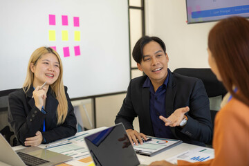 Three people are sitting at a table in a conference room, discussing business matters. The man in the middle is wearing a suit and he is the leader of the group. The woman on the left is smiling
