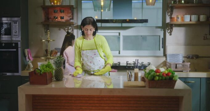 Indian mom standing wear apron and hand gloves collect pick up waste trash in box day time adult woman lady do work teen age girl child use rag wipe table dust keep fresh neat tidy clean kitchen home