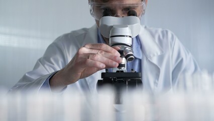 Male scientist wearing lab coat and protective glasses using microscope doing research in modern laboratory setup, portrait view. Low key lighting photo. Science and Medicine concept