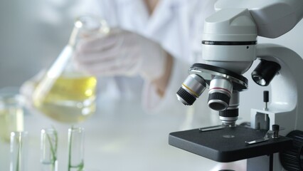 A scientist, wearing a lab coat and pink protective gloves, is pouring a yellow oily liquid from one beaker to another near microscope in laboratory, close up. Medicine and science