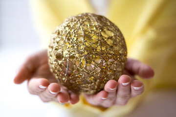 Gentle Hands Cradling Intricate Golden Christmas Ornament Against Soft Yellow Background