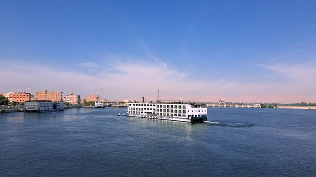 Multiple white cruise ships glide on the blue Nile River, under a clear blue sky, near Esna Lock, Egypt. The distant cityscape and calm waters create a picturesque travel scene.