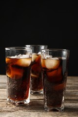 Refreshing cola with ice cubes in glasses on wooden table against black background, closeup