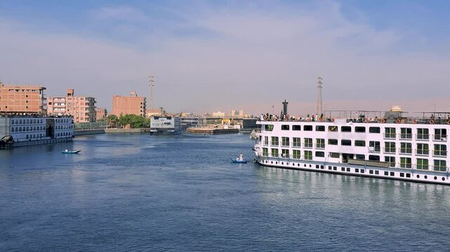 Multiple white cruise ships glide on the blue Nile River, under a clear blue sky, near Esna Lock, Egypt. The distant cityscape and calm waters create a picturesque travel scene.