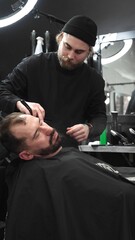 Professional barber carefully shaving client's beard with straight razor, applying gel and using...