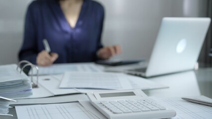 Magnifying glass, white calculator, pen and documents are lying on accountant's desk. Businesswoman wearing blue dress is working with documents on the background. Business and audit concept