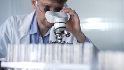 Male scientist wearing lab coat and protective glasses using microscope doing research in modern...