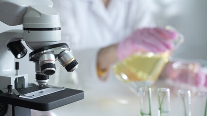 A scientist, wearing a lab coat and pink protective gloves, is pouring a yellow oily liquid from one beaker to another near microscope in laboratory, close up. Medicine and science