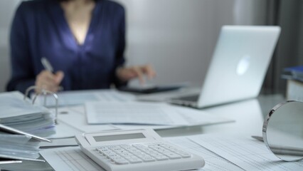Magnifying glass, white calculator, pen and documents are lying on accountant's desk. Businesswoman wearing blue dress is working with documents on the background. Business and audit concept
